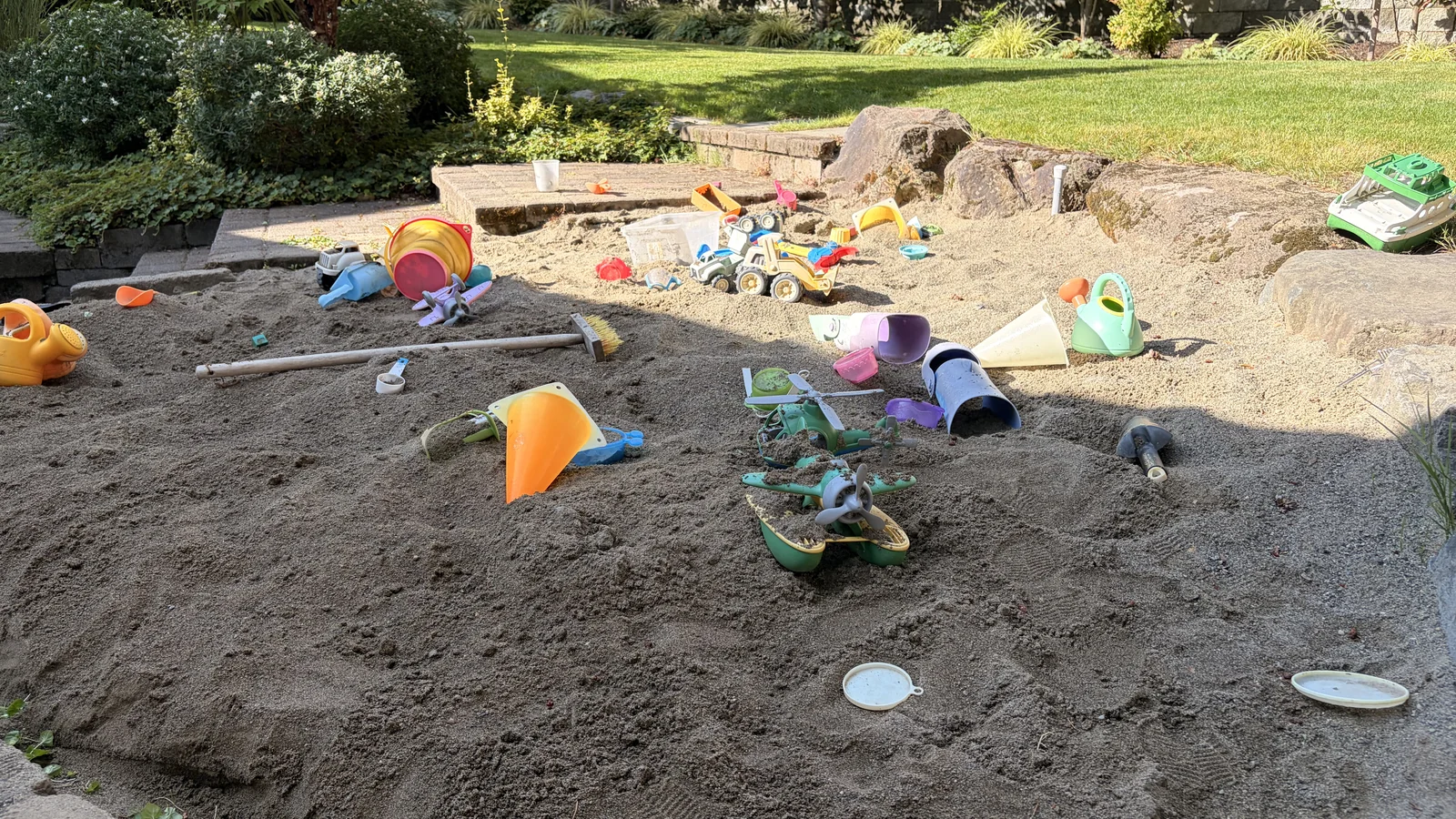 Backyard sandbox filled with light sand and scattered toys, bordered by rocks with the lawn sloping up behind it