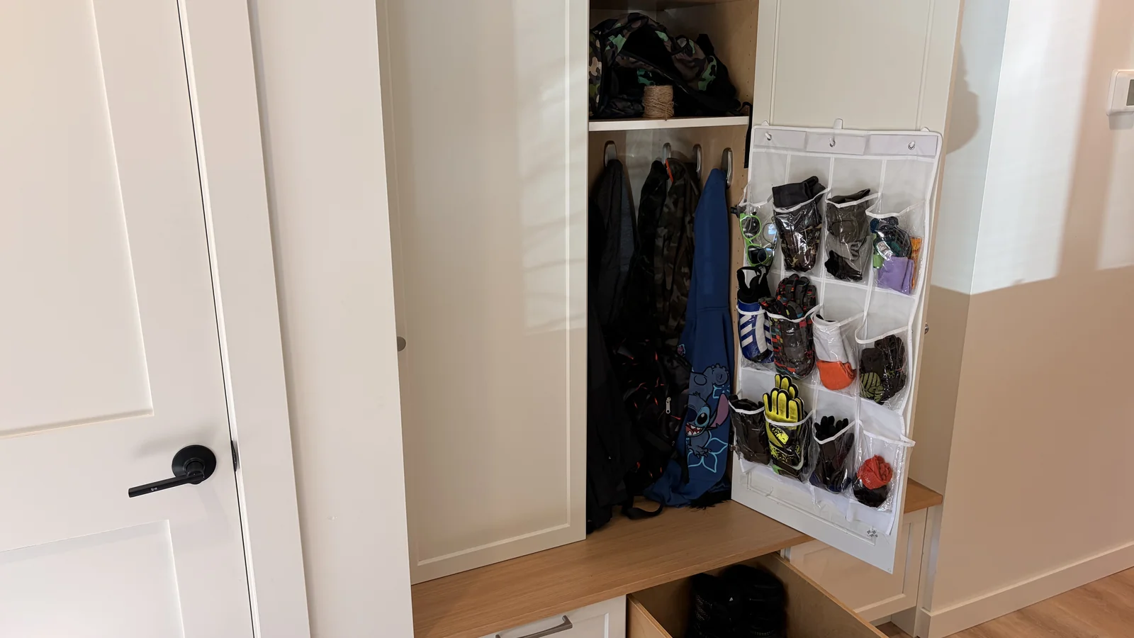 Open built-in entry cubby with coats on hooks, a hanging door organizer for gloves, and a shoe drawer pulled out under the bench