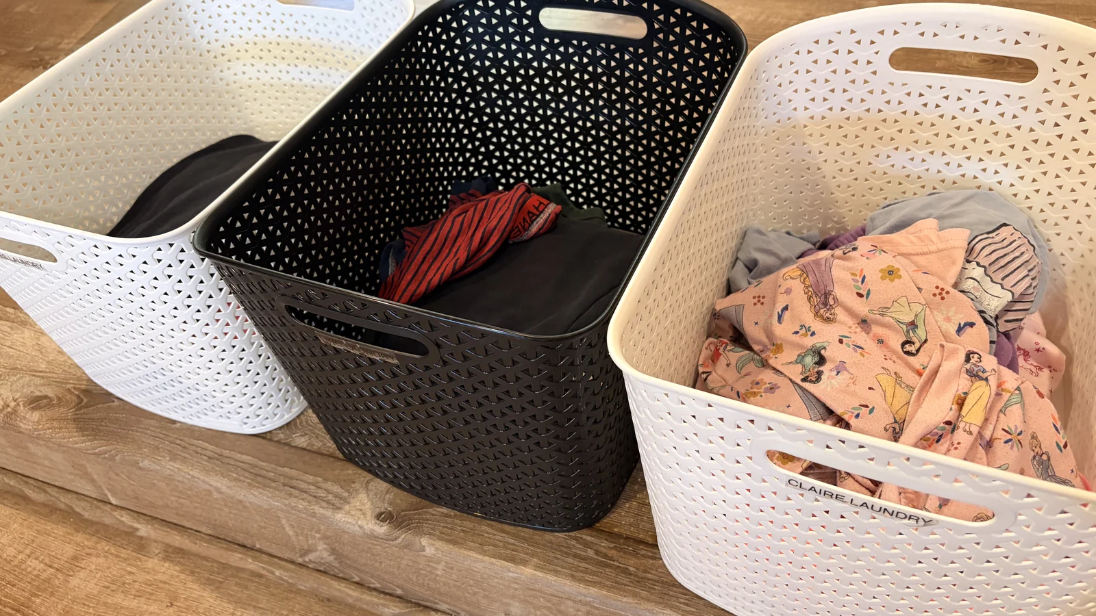 Three named plastic laundry bins lined up with unfolded clean clothes sorted inside