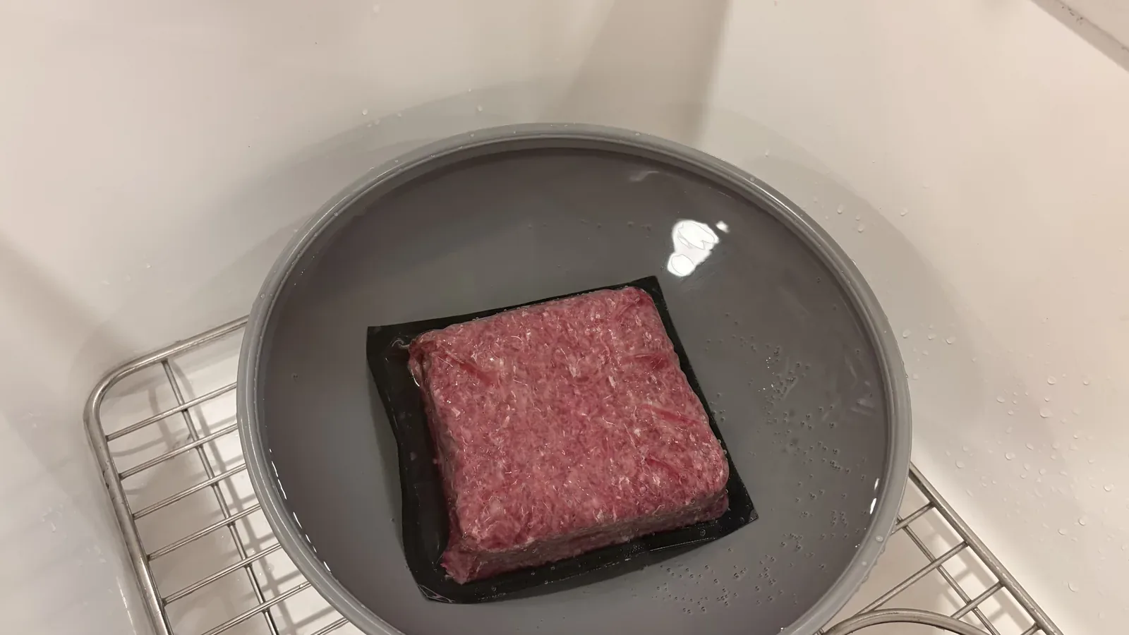 Vacuum-sealed ground beef thawing in a grey bowl of water in the kitchen sink.